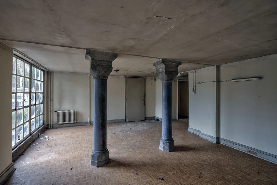 Sala vacía de parqué con dos columnas oscuras y puertas acristaladas izquierdas.