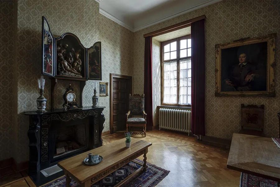 Wohnraum mit dunklem Kamin, Tisch und Stühlen an einem hohen Fenster sowie Porträts an der Wand.