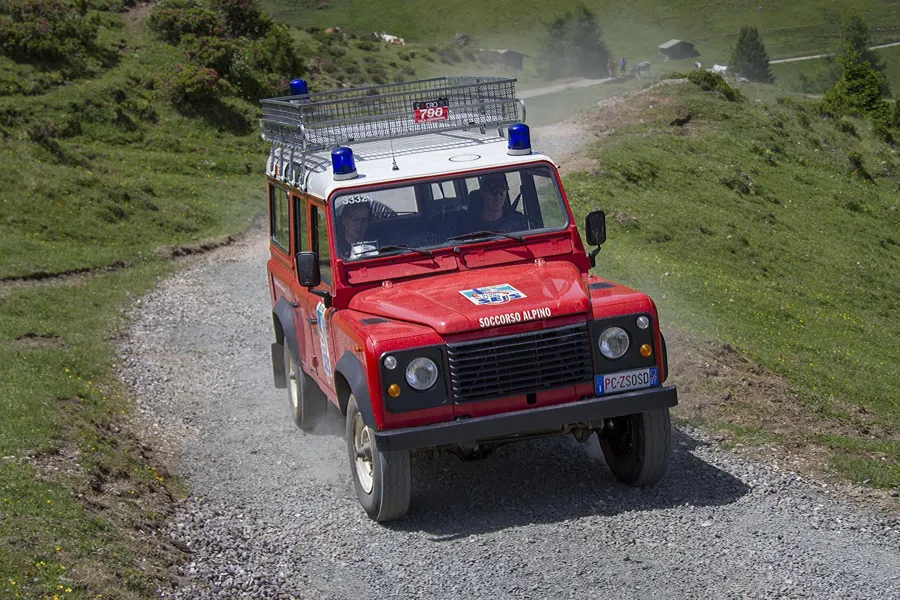 Todoterreno rojo de rescate de montaña por una pista de grava entre prados alpinos verdes.