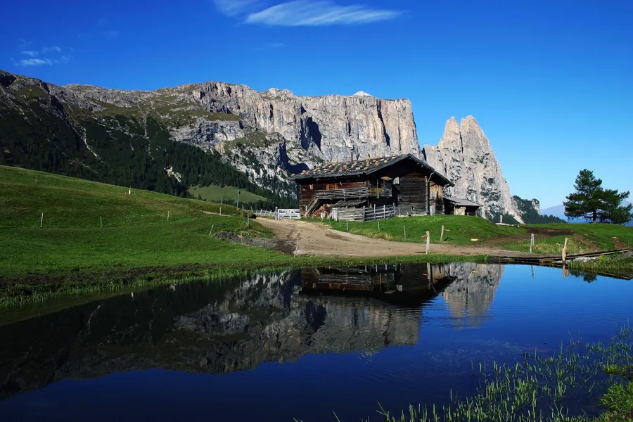 Cabaña de madera y pared escarpada del Schlern reflejadas en una pequeña laguna alpina.