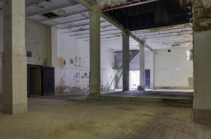 Leere geflieste Werkshalle mit quadratischen Säulen, Treppe und abblätternder Decke.
