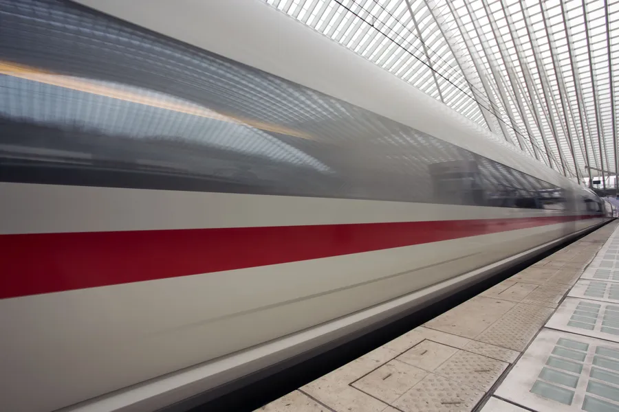 Tren blanco con franja roja como una larga estela de movimiento junto al andén bajo el techo de Lieja-Guillemins.