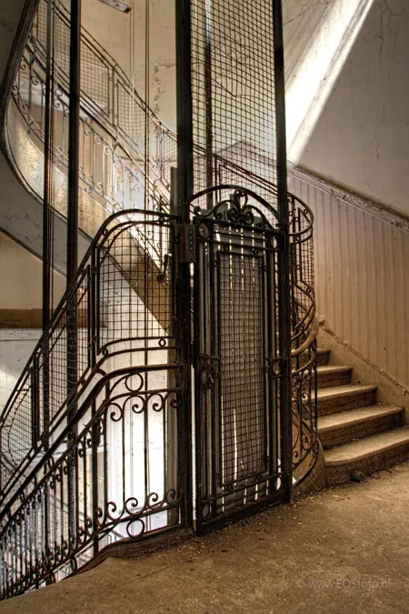 Ancienne cage d’ascenseur à grille ouvragée entre deux volées d’escalier.