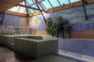 Green-tiled round pool beneath a glass roof beside a colourful mosaic wall.