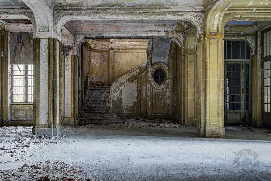 Leere Halle mit gelben Ziersäulen und geschwungener Treppe hinten.