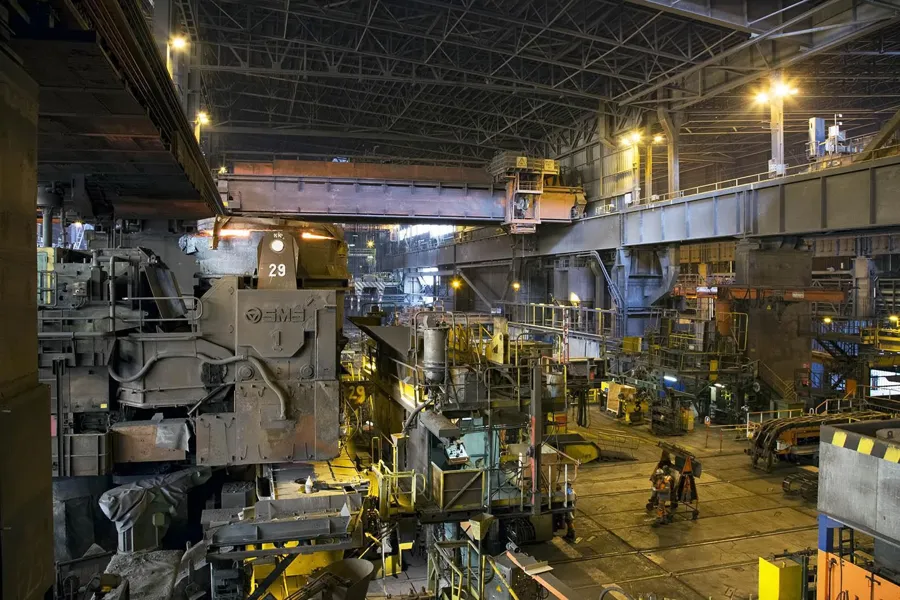 Grand hall sidérurgique avec machines lourdes, passerelles jaunes et pont roulant sous le toit métallique.