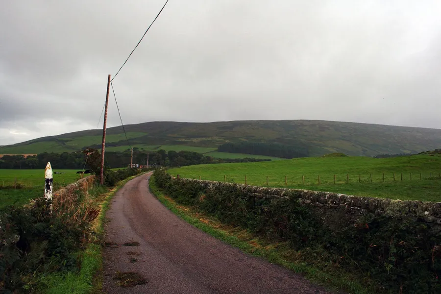 Schmale Straße zwischen niedrigen Steinmauern und grünen Feldern bei Mull of Kintyre unter grauem Himmel.