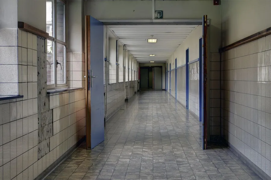 Pasillo largo de hospital alicatado con ventanas a la izquierda y marcos de puertas azules.