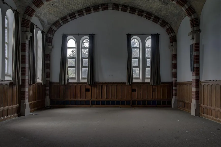 Sala vacía con revestimiento de madera y ventanas altas bajo arcos a franjas.