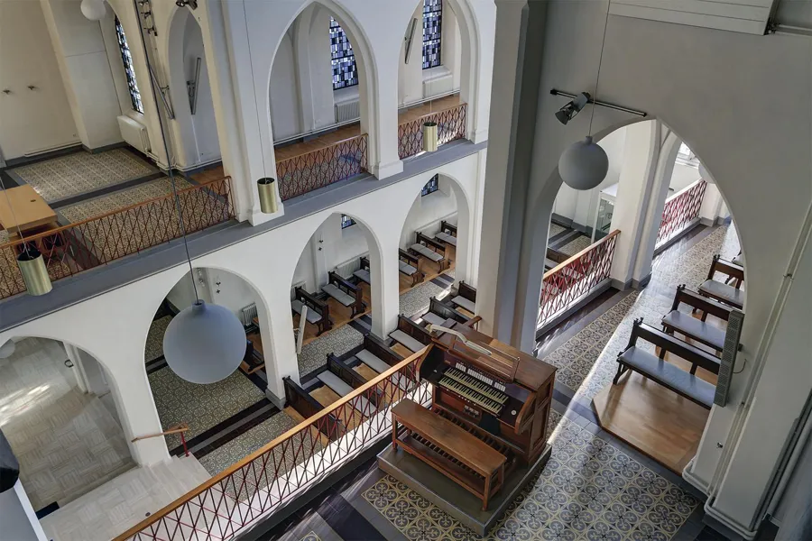 Hoog schuin uitzicht vanaf de galerij op kapelbanken, balustrades en hanglampen.