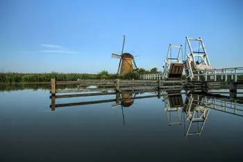 Windmill and white wooden drawbridge reflected in still water beneath a clear blue sky.