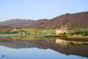 Eilean Donan Castle en de stenen toegangsbrug weerspiegelen in rustig water, met heuvels op de achtergrond.