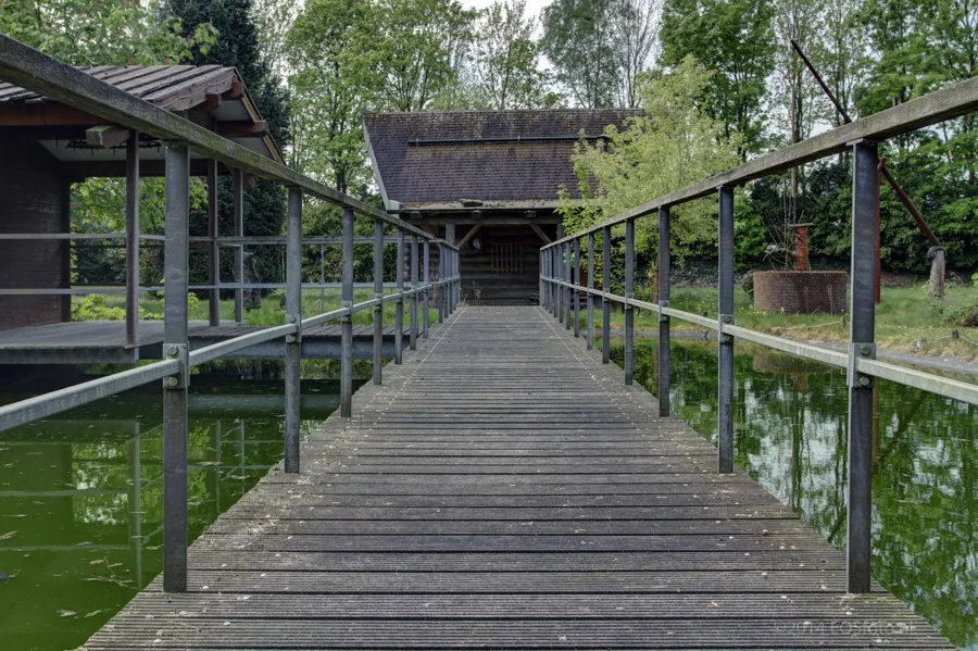 Puente de madera con barandillas metálicas sobre agua verde hacia una cabaña de sauna.