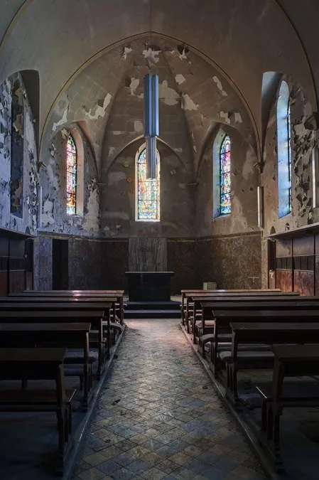 Donkerder zicht door de beschadigde kapel naar het altaar en drie hoge ramen.