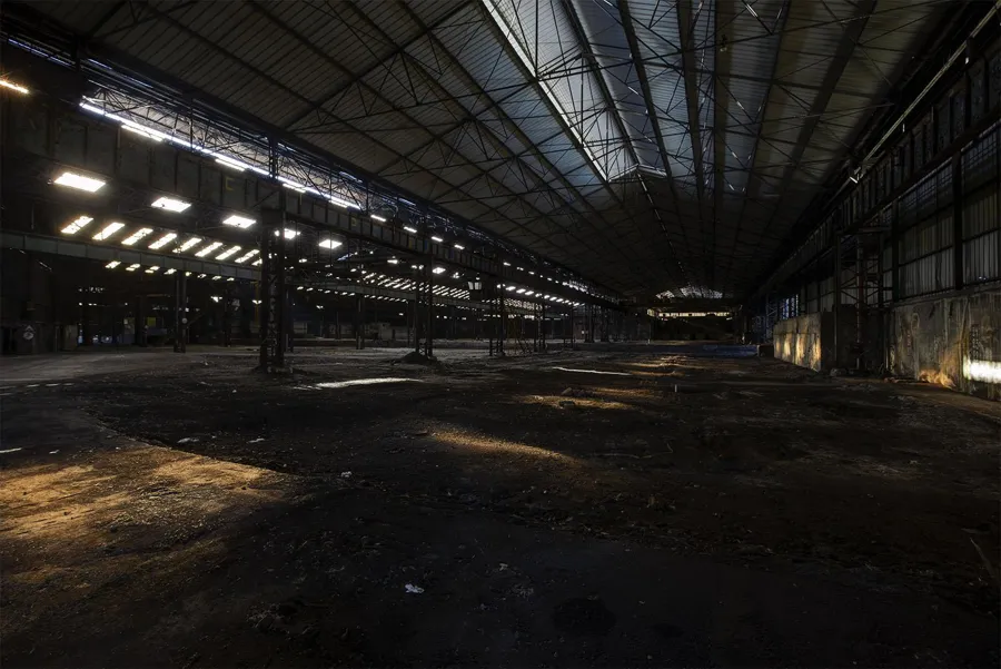 Grande halle industrielle vide aux taches de lumière sous un toit d’acier.