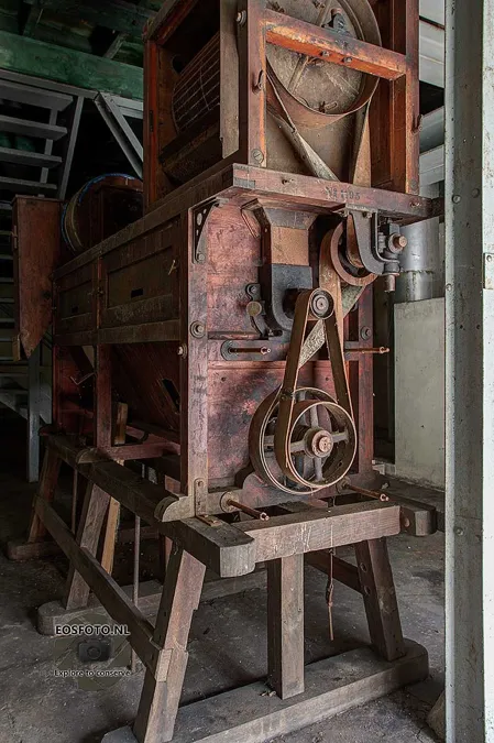 Staande opname van een hoge houten machine met ronde riemschijven en een smalle trap ernaast.
