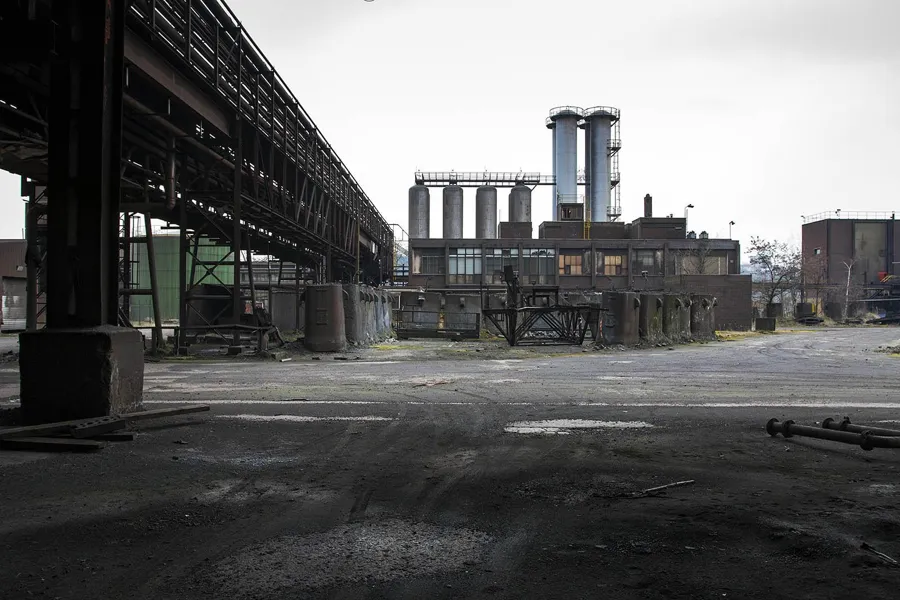 Wide abandoned yard beneath an elevated conveyor structure near chimneys.