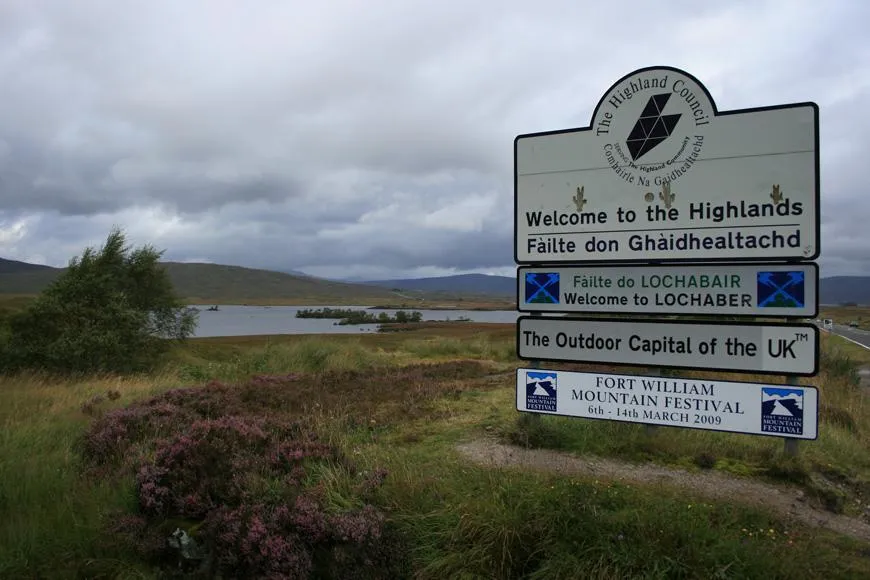 Willkommensschild für die Highlands und Lochaber an einer Straße, mit Heide und See im Hintergrund.