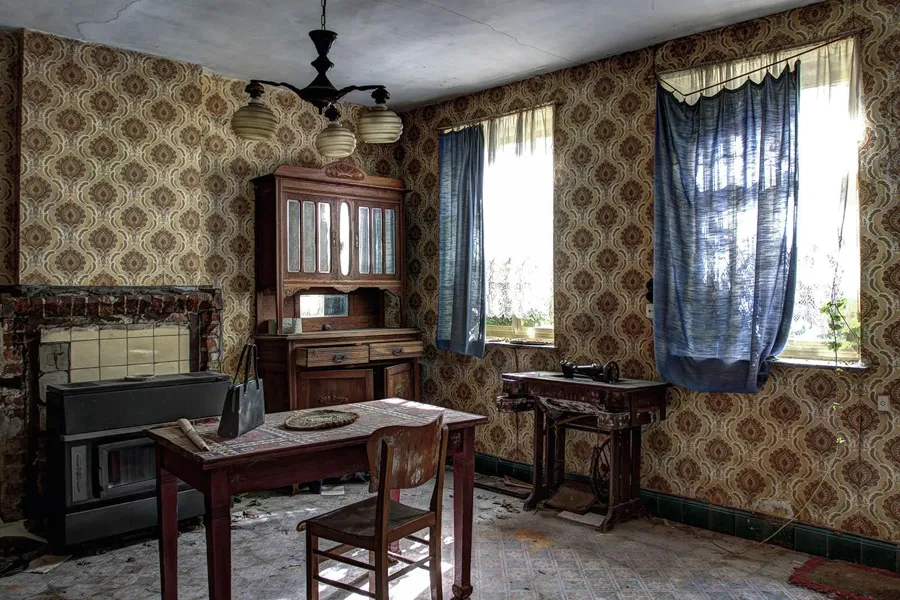 Table and chair beside two curtained windows in a room with patterned wallpaper
