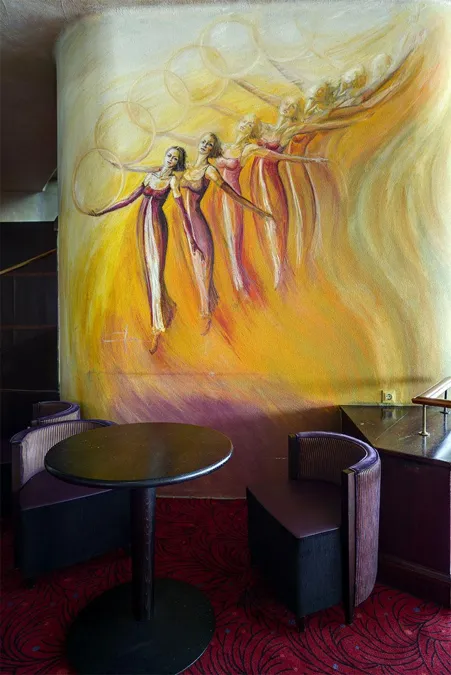 Mural amarillo con figuras bailando sobre dos mesitas redondas y sillones oscuros.
