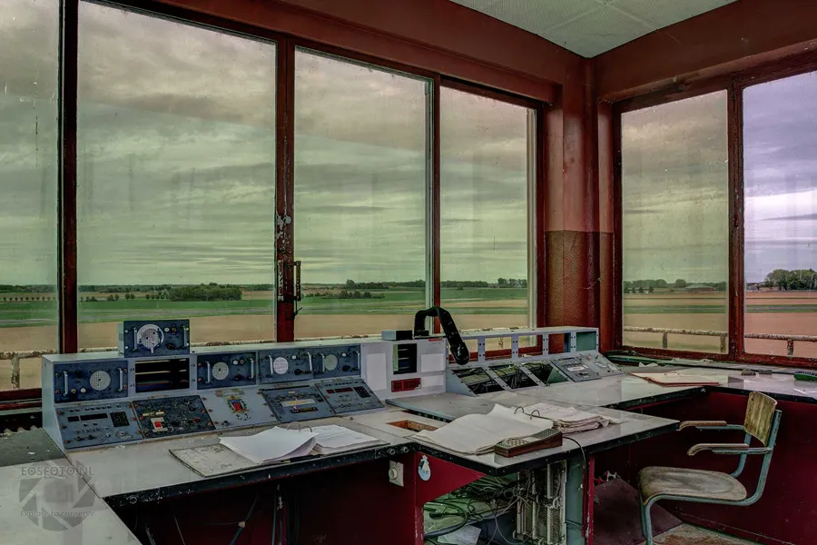 Old control consoles beside large windows overlooking a flat grassy field.