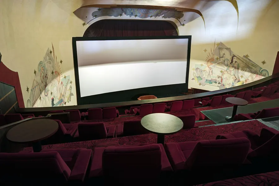 Pantalla de cine desde el balcón, con sillones rojos, mesitas redondas y murales alrededor.