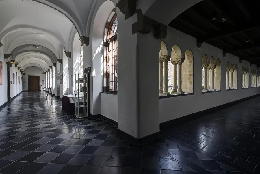 Ecke zweier Klostergänge mit dunklen Bodenfliesen, weißen Gewölben und Sonnenlicht durch hohe Fenster.