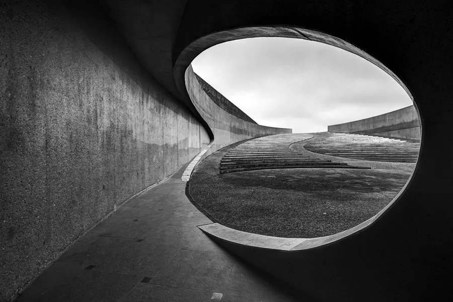 Zwart-witte doorkijk door een ronde betonnen opening naar een brede gebogen buitentrap onder een bewolkte lucht.