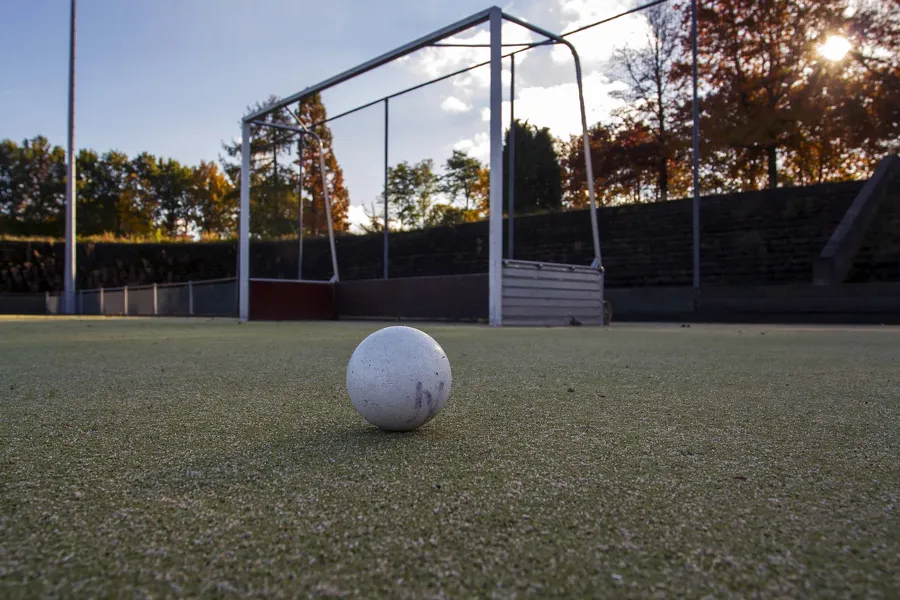 Bola blanca de hockey a ras del césped artificial, con portería y gradas oscuras al fondo.