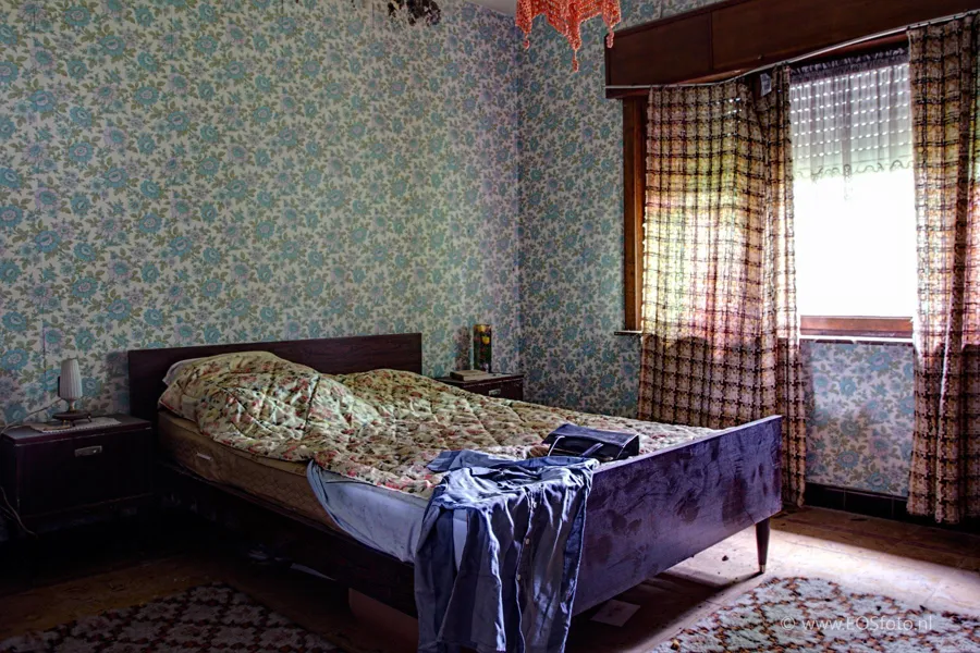 Wooden bed beside a window with checked curtains in a bedroom with floral wallpaper