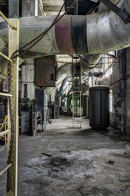 Long factory passage beneath thick pipes beside a black tank.
