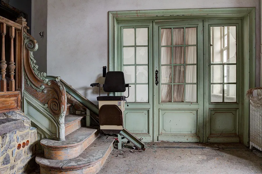 Stair foot with an ornate banister and a chair beside green double doors.