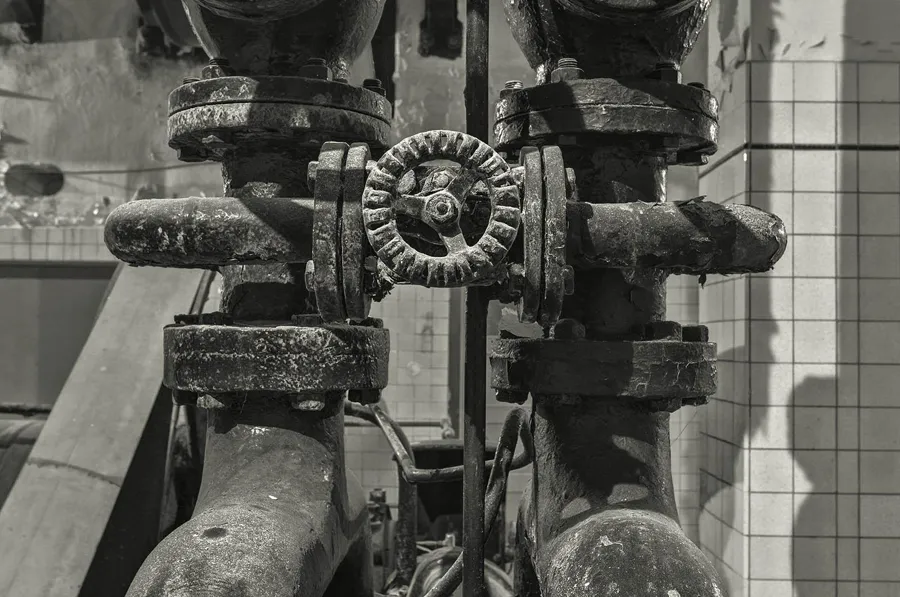Schwarz-Weiß-Detail eines kleinen Zahnrads zwischen dicken Rohren und großen Verschraubungen.