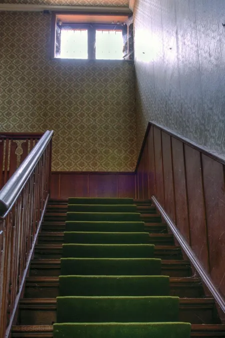 Escalera con moqueta verde hacia un rellano con una pequeña ventana