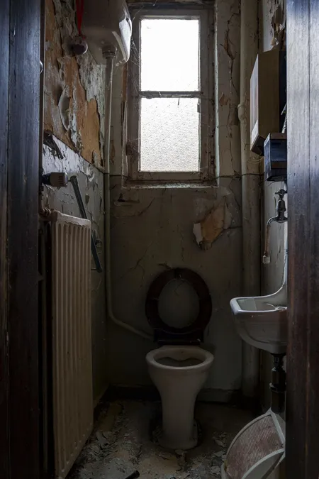 Smalle vervallen toiletruimte met hoog raam, wc en kleine wastafel.