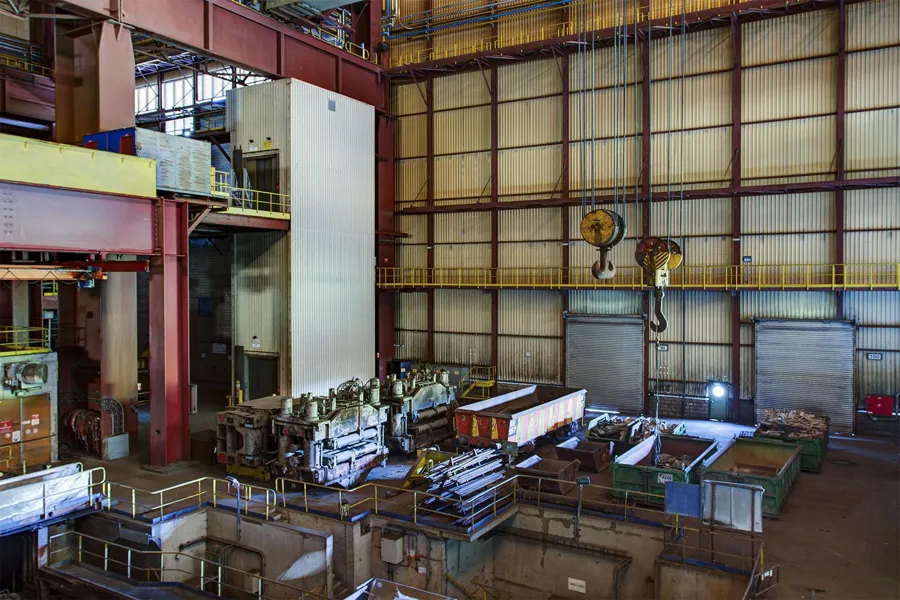 Tall bright factory hall with crane hooks and numerous loose machine parts.