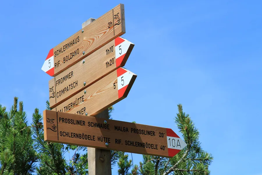 Señales de senderismo de madera con puntas rojas y blancas y nombres de rutas contra cielo azul.