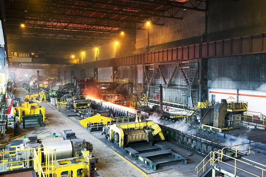 Longue bande d’acier orange dans un haut atelier, avec machines jaunes et brume au fond.