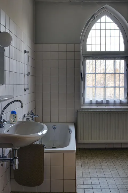 Kleine betegelde badkamer met wastafel, bad en hoog spitsboograam.