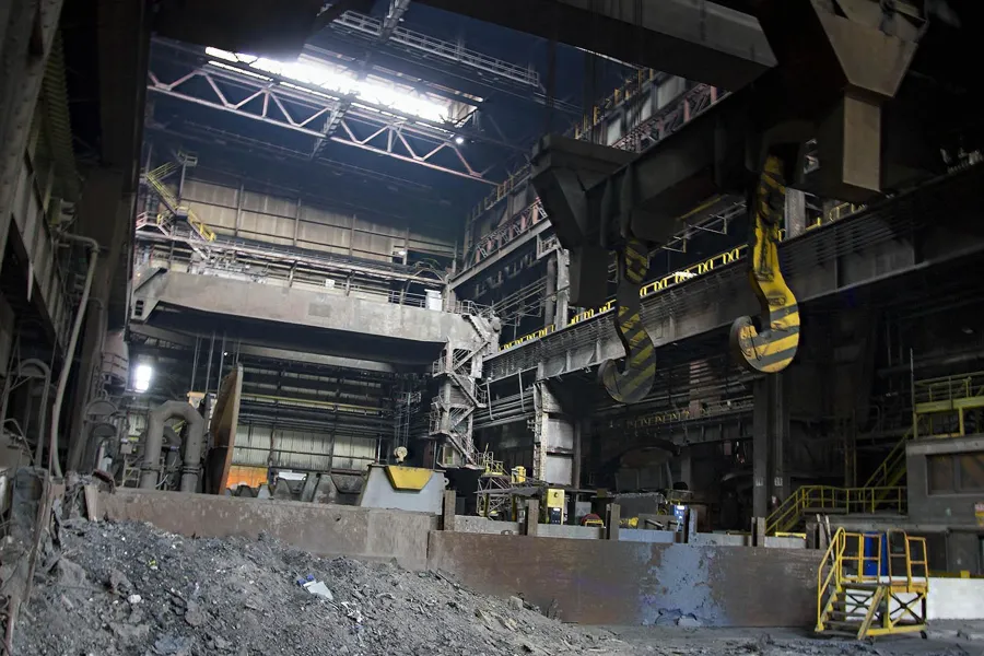 Tall steelworks hall with yellow crane hooks and rubble on the floor.