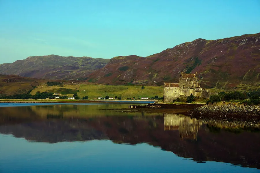 Eilean Donan Castle und seine steinerne Zugangsbrücke spiegeln sich in ruhigem Wasser vor Hügeln.