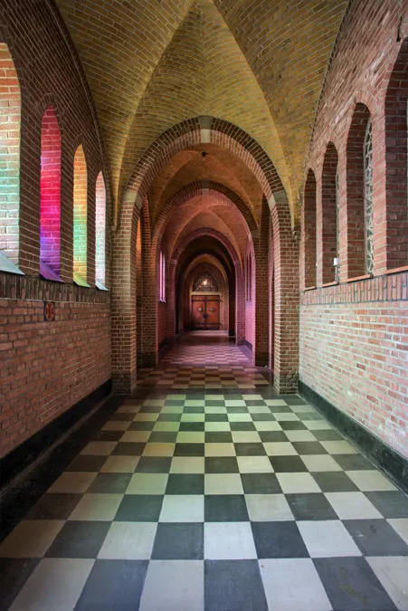 Luz de ventanas coloreadas sobre muros de ladrillo de un pasillo monástico.