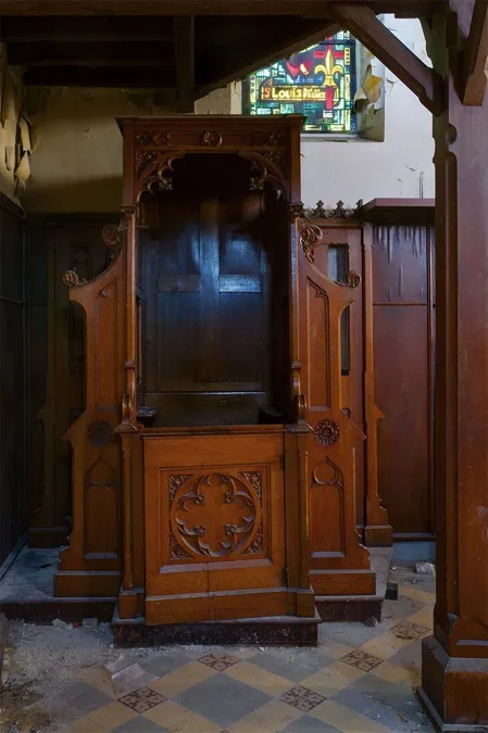 Houten biechtstoel met snijwerk onder een klein gekleurd raam.