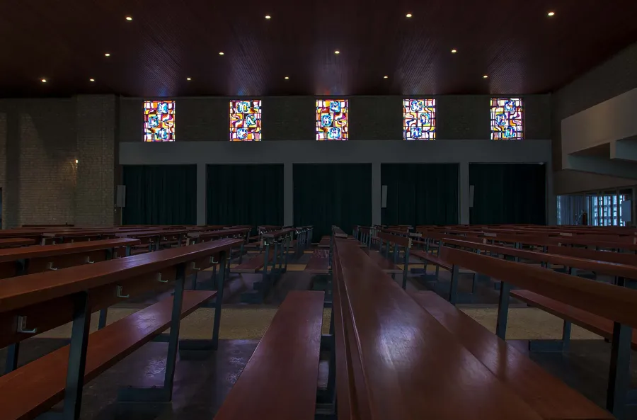 Long wooden pews beneath a dark ceiling with five small colourful windows behind.