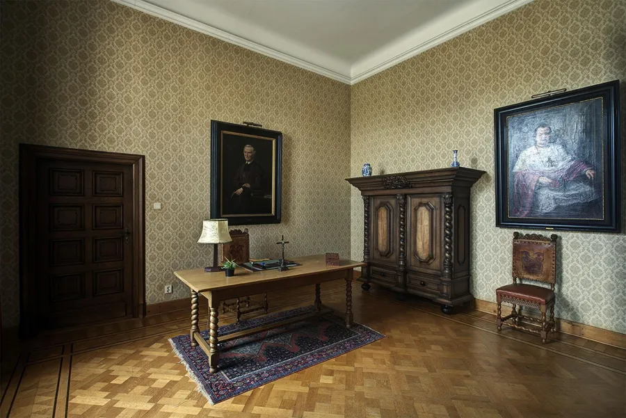 Historisches Zimmer mit Schreibtisch auf einem Teppich, Holzschrank und zwei gemalten Porträts.