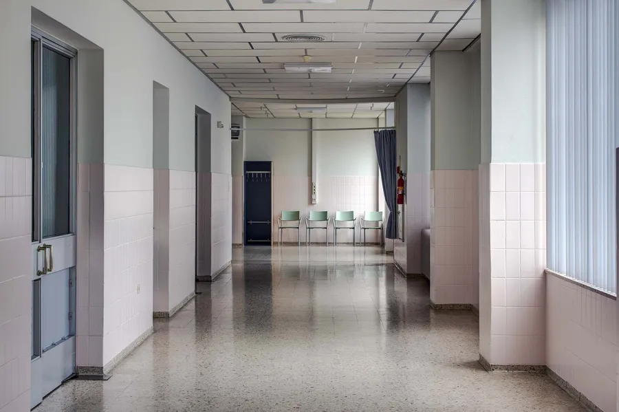 Leerer Krankenhausflur mit hellen Wänden, Türen links und Tageslicht durch hohe Fenster rechts.