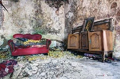 Sofá rojo y armario de madera en una habitación con paredes desconchadas y escombros en el suelo