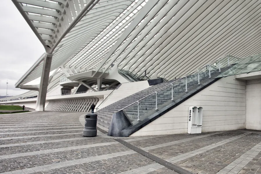 Escalera ancha junto a una pared blanca de hormigón, con una papelera y nervaduras inclinadas encima.