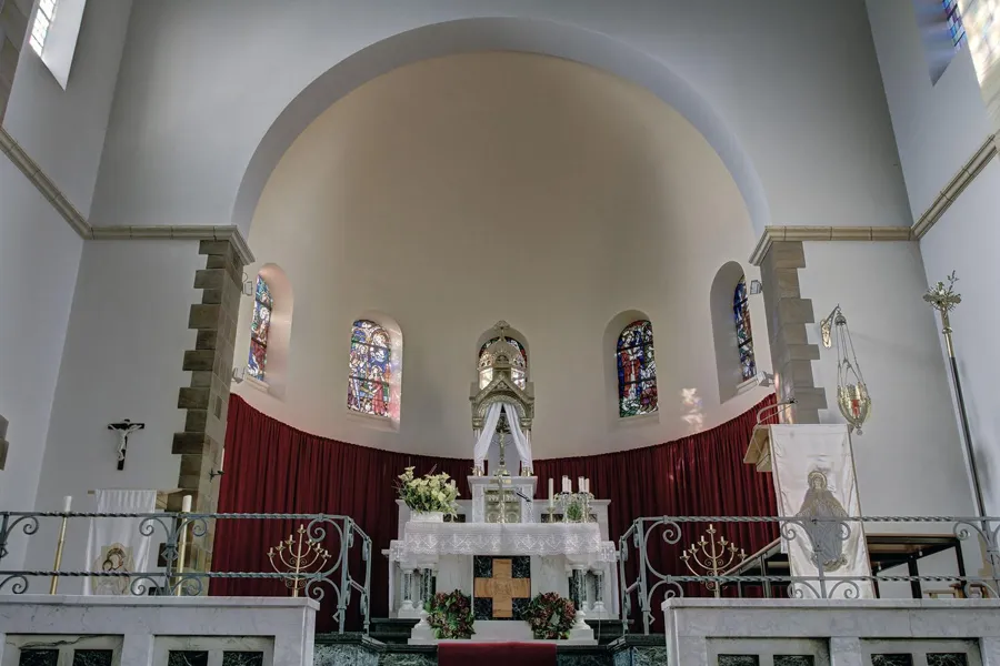Vista baja hacia el altar blanco y la pared curva del presbiterio, con vidrieras sobre cortinas rojas.