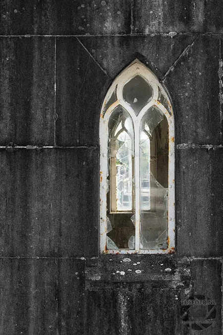 Schmales Spitzbogenfenster in dunkler verwitterter Kapellenwand.